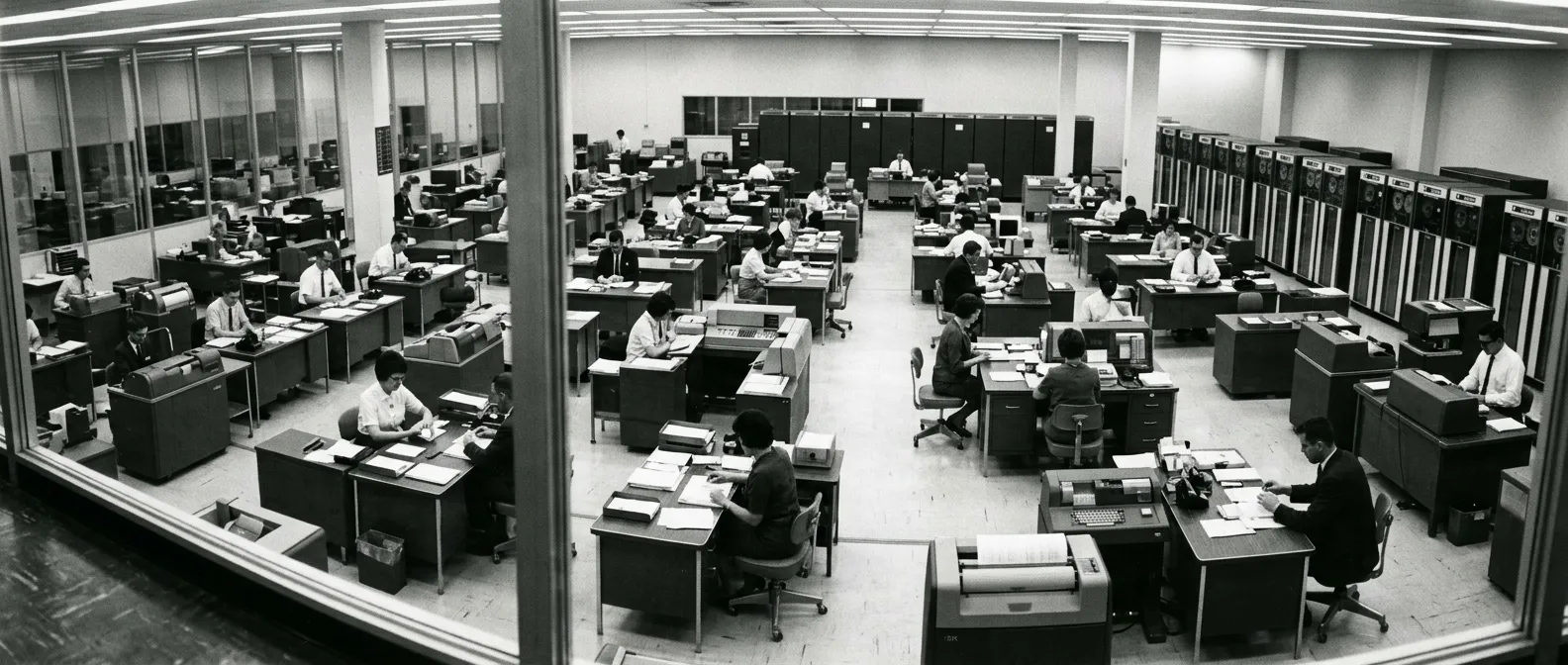 A data-processing floor seen from a glass-fronted office above it: rows of desks, an operator at each with a terminal or a card sorter, tape cabinets along the far wall.