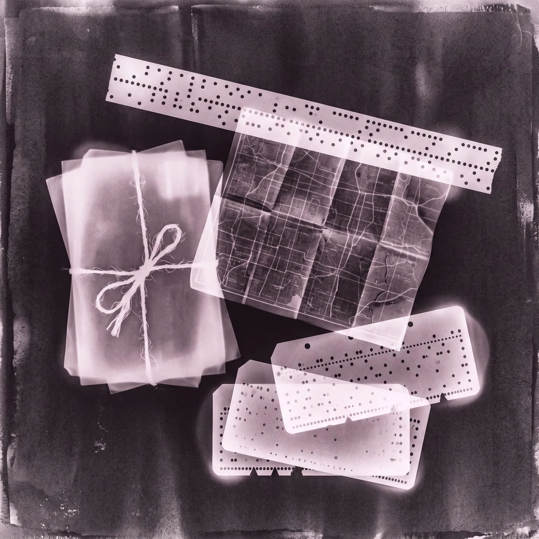 A photogram of a bundle of letters, a folded map, punched tape and cards, toned plum-grey.