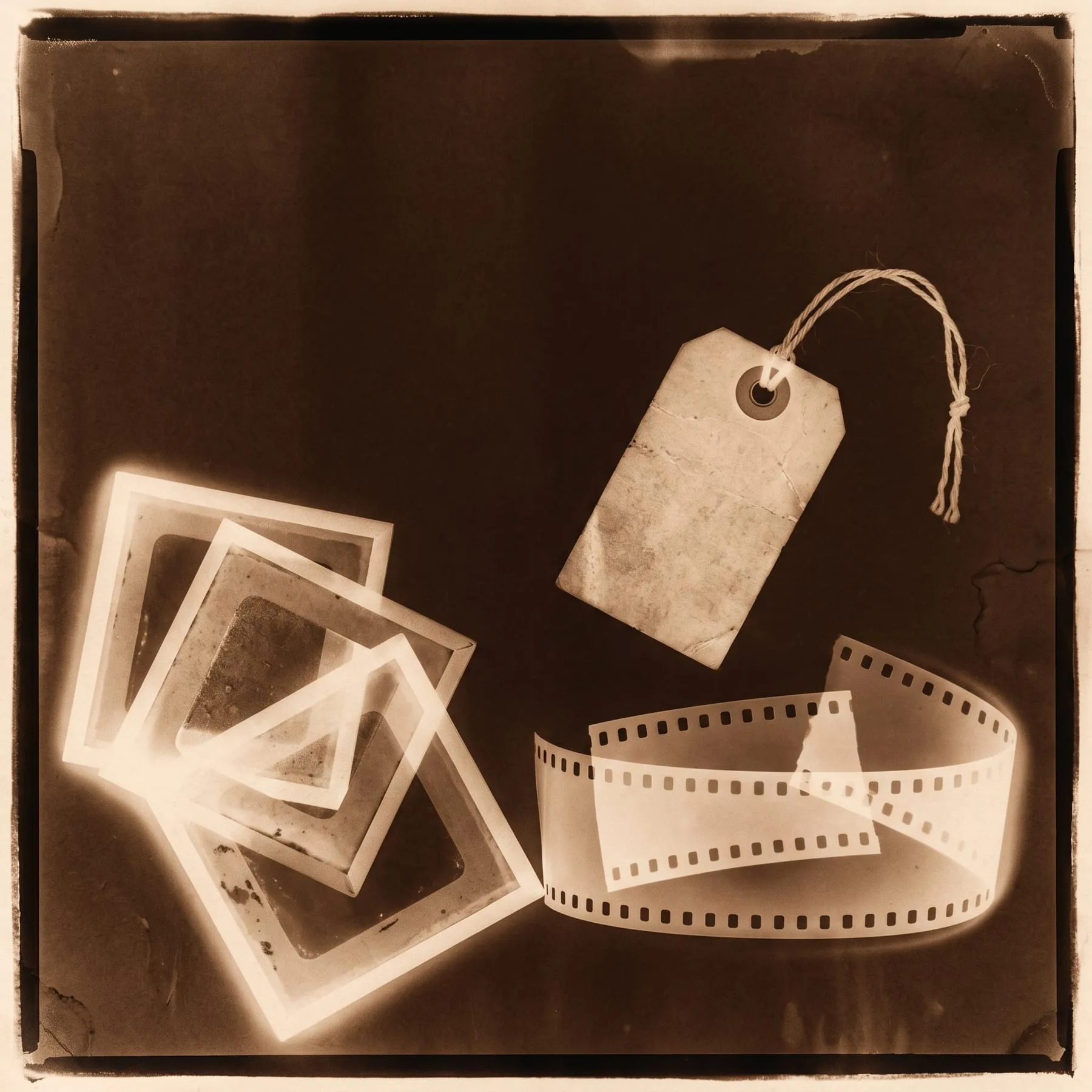 A photogram of three lantern slides fanned from one corner, a strip of film and a luggage tag, toned sepia.